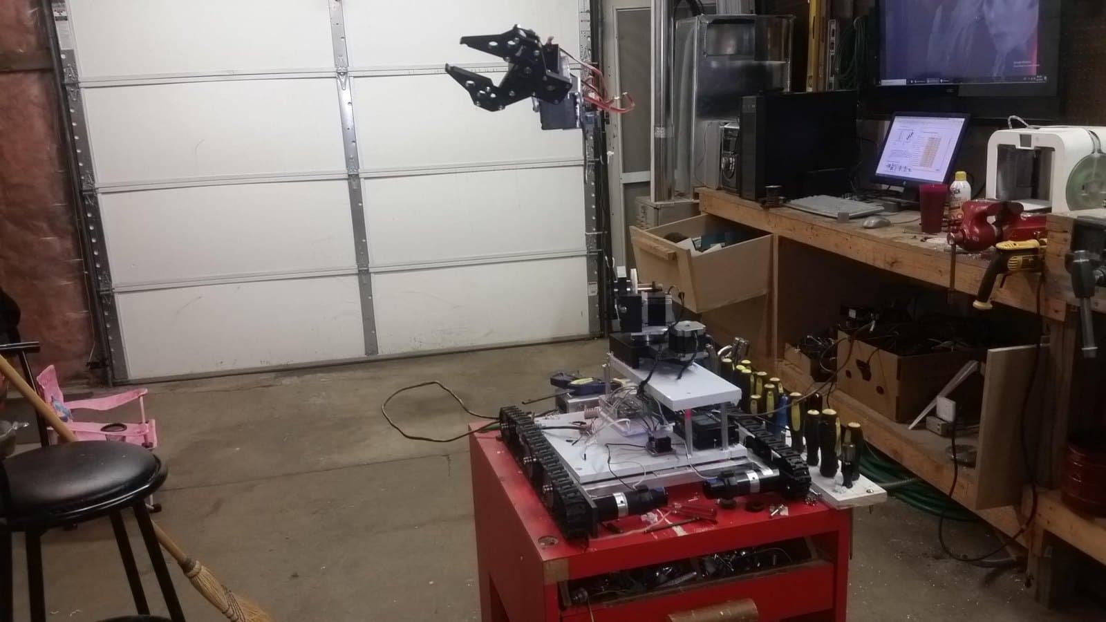 Arm raised, claw open; the shop behind it with the PC, the TV and the 3D printer