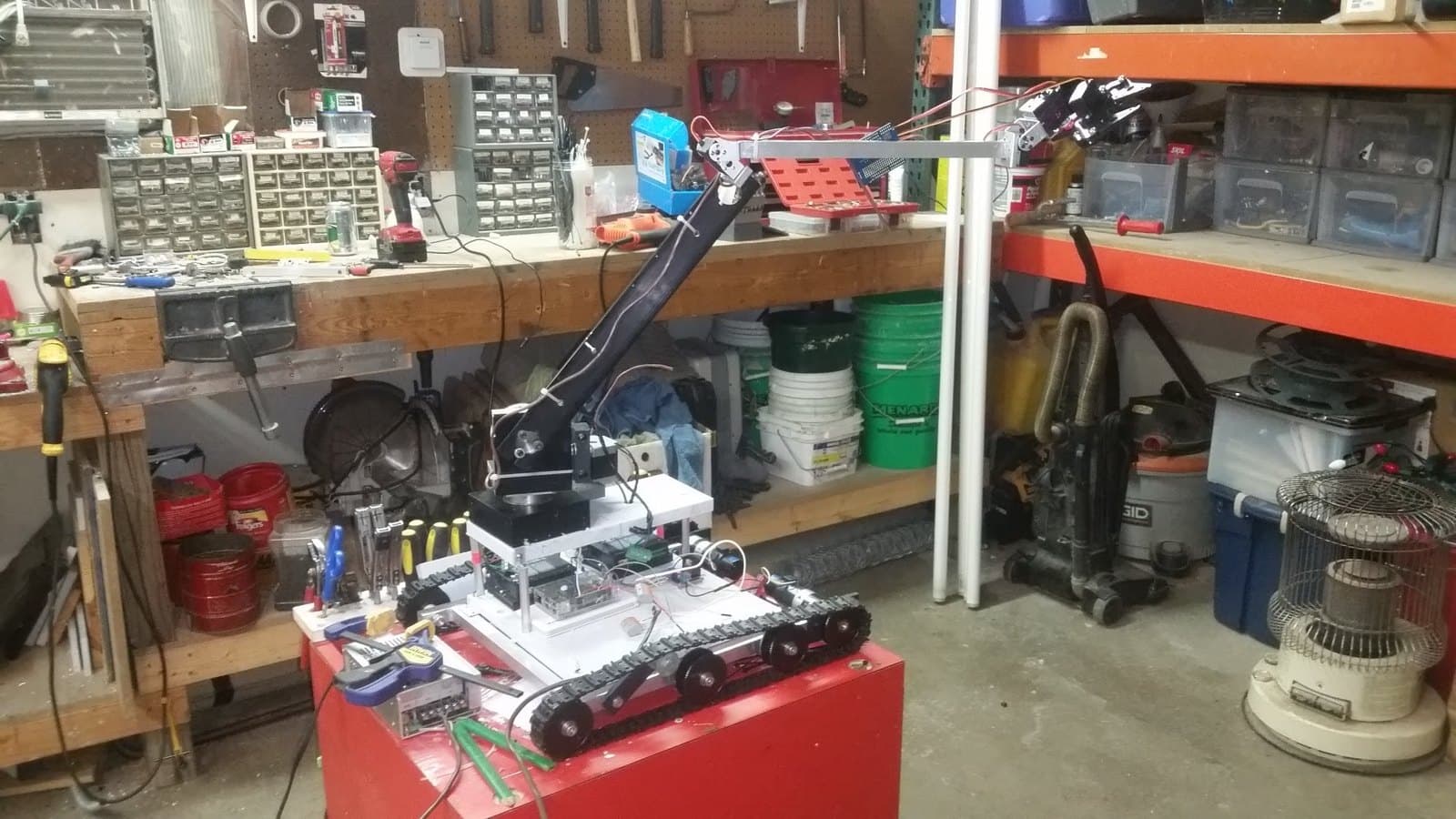 Tracked robot on a red shop cart, arm extended over the workbench